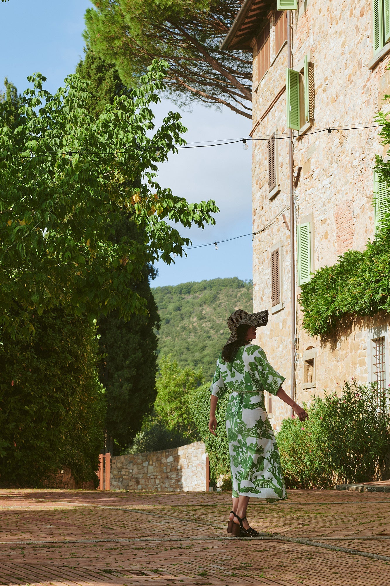 Woman photographed from behind walking outdoors wearing green willow print dress and large sun hat