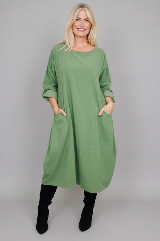 A woman wearing a loose sage green long-sleeve tunic dress with front patch pockets, paired with black boots, standing against a white paneled wall.
