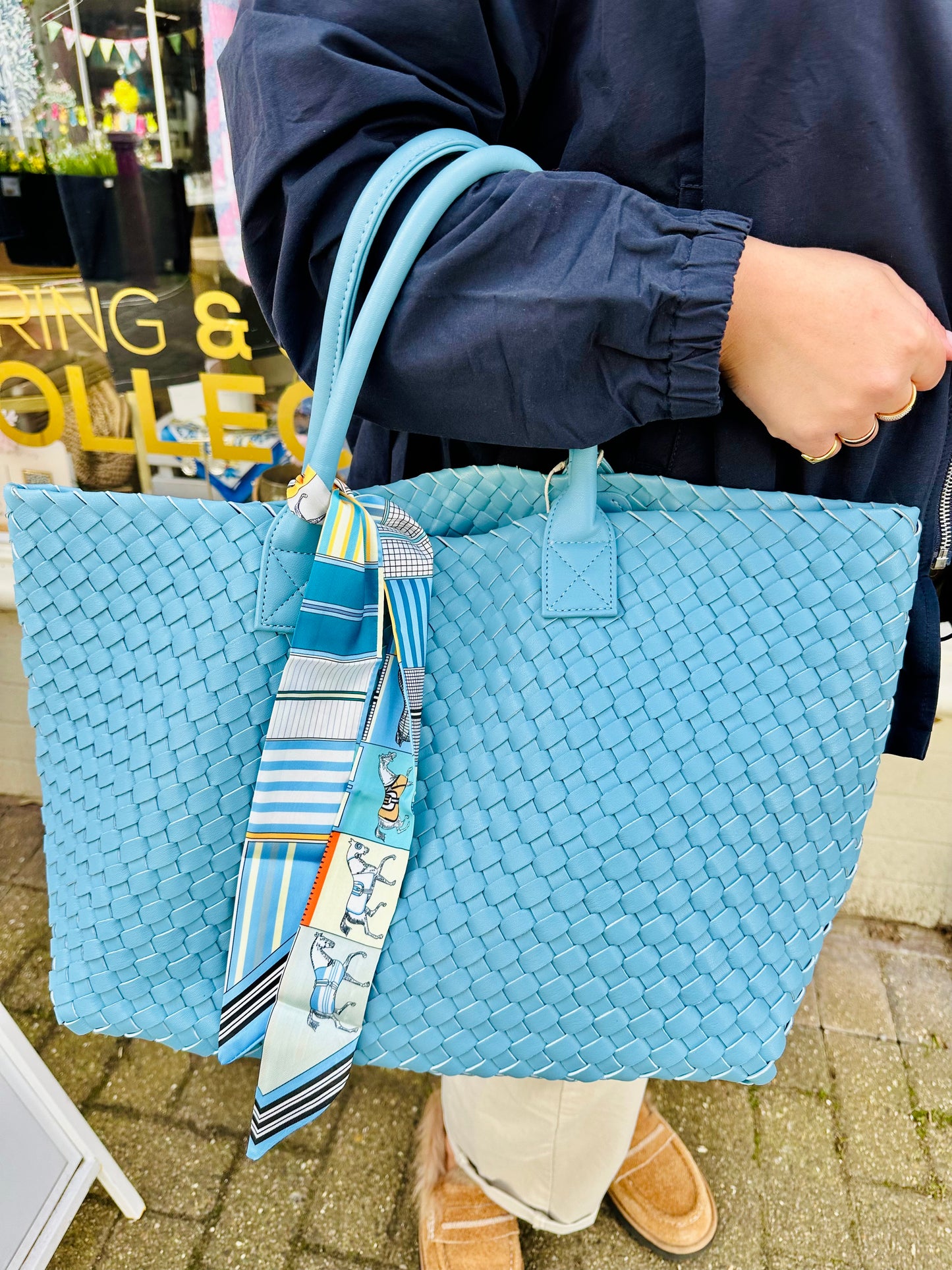 Blue woven tote handbag with double rounded handles and a decorative printed scarf attached, held by a person wearing cream trousers and tan footwear.